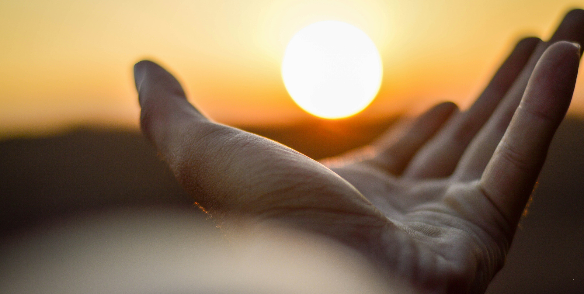 Bilde av en hånd med håndflaten opp, som strekker seg mot solen (en hvit kugle), i horisonten. Pga avstand ser det ut som at hånden er i ferd med å omfavne solen. Copyright Aaamir Suhail on Unsplash