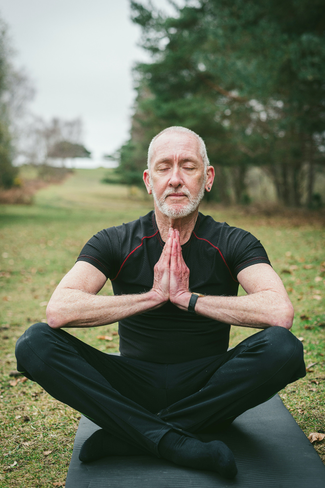 Copyright Margaret Jaszowska on Unsplash. Bildet viser en eldre mann med grått hår og skjegg, kledd i sort , sittende i lotus på gressplenen, med lukkede øyne og hendene i namaste. 