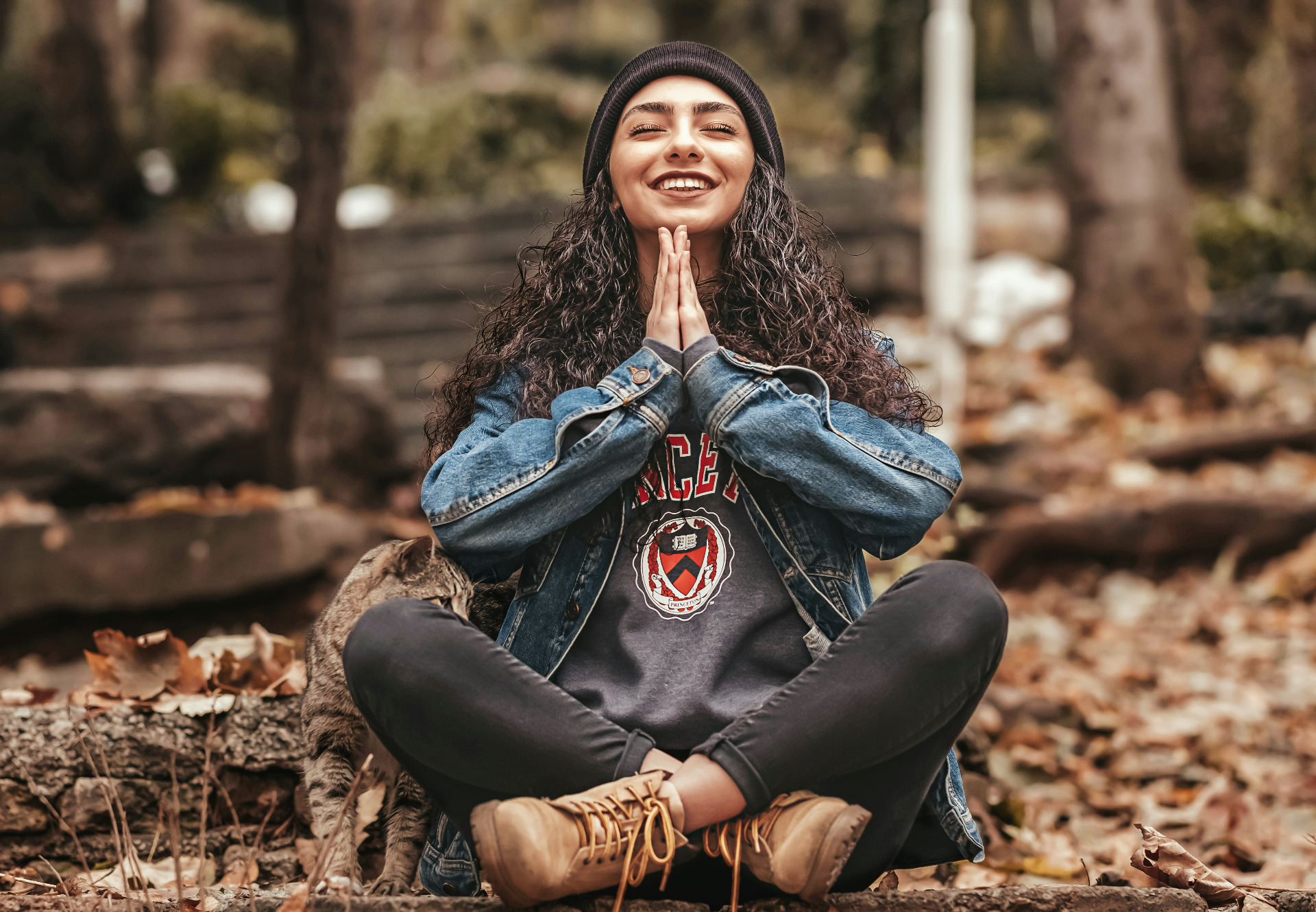 Copyright Omid Armin on Unsplash. Bildet viser en ung kvinne som sitter i skredderstilling på jorden dekket med løv. Kvinnen har hendene i namaste under haken og smiler lykkelig med lukkede øyne.