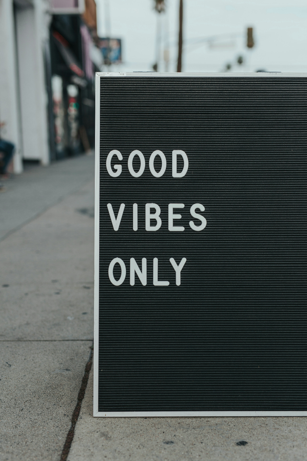 Copyright Mark Adriane on Unsplash. Bildet viser et skilt som står på grå fliser på en gate (sløret bakgrunn). På skiltet står det hvit på sort "Good Vibes only"